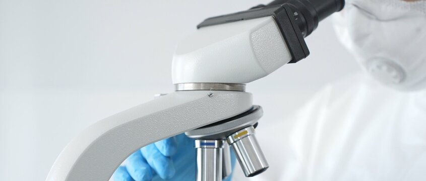 Medical virologist in a hazmat suit, mask, and blue gloves, diligently working at a microscope in a laboratory researching viruses. Healthcare, medicine, and science concept