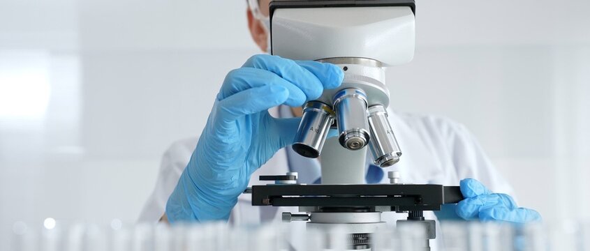 Woman science researcher wearing protective gear is checking microscope, analyzing blue test tubes in modern laboratory research setting. Medicine, and science concept