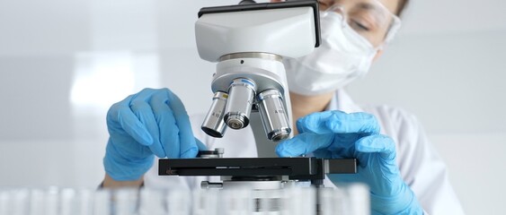 Woman science researcher wearing protective gear is checking microscope, analyzing blue test tubes...