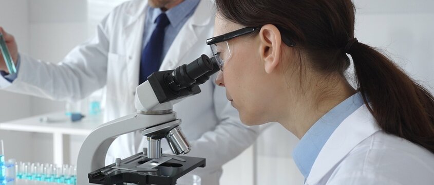Female scientist wearing protective glasses looking through a microscope during medical research in laboratory with her male colleague working in the background. Medicine and science concept