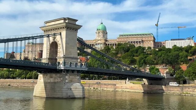 While floating under the Budapest Chain Bridge (aka Bridge of Pearls), the camera points at the magnificent Buda Castle high on a hill in the background, on a beautiful summer day in Hungary.