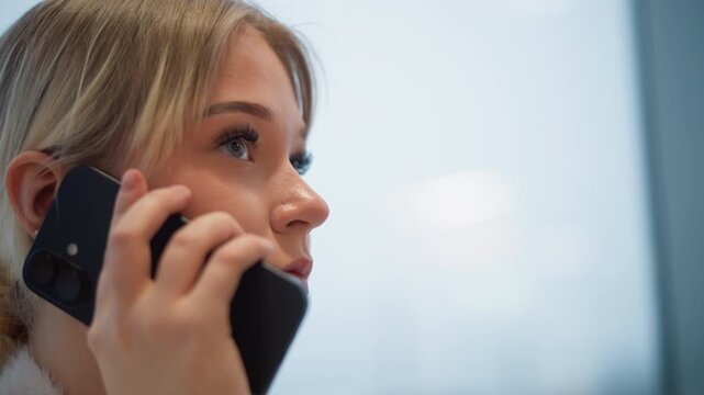 Head-and-shoulders side profile of young woman holding smartphone near ear while speaking, with focused expression, steady grip, soft indoor light, and blurred background suggesting active call