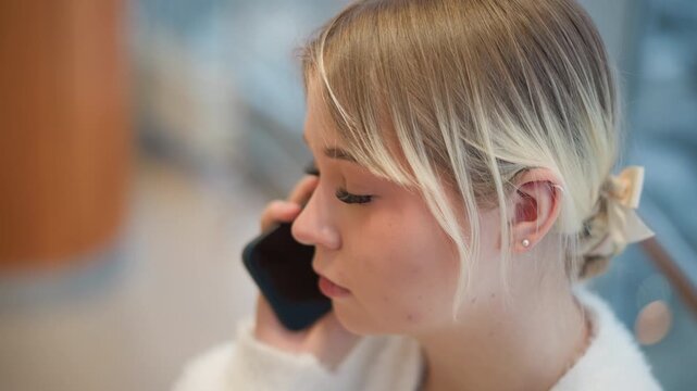 Close profile of young blonde woman holding smartphone to ear, engaged in conversation with attentive, slightly concerned expression, subtle head tilt, and soft indoor background, with woman, phone