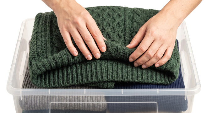 Hands folding a warm green cable knit sweater into a clear storage bin