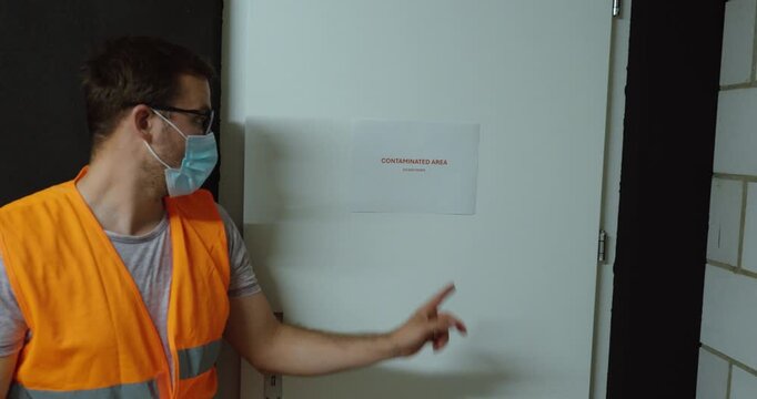 Worker wearing a face mask installs a Contaminated Area warning sign on a door. Ideal for contamination control, restricted access and workplace safety.