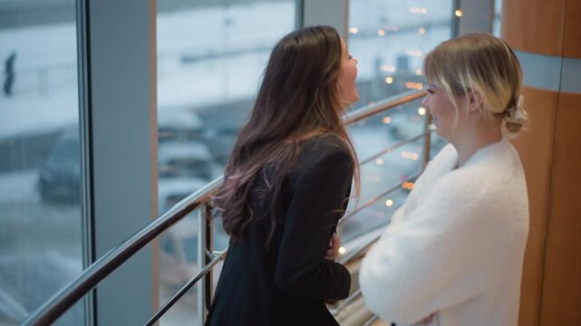Two women stand near bright atrium windows and railing inside modern workspace, sharing friendly conversation with expressive gestures, eye contact, relaxed posture, natural daylight