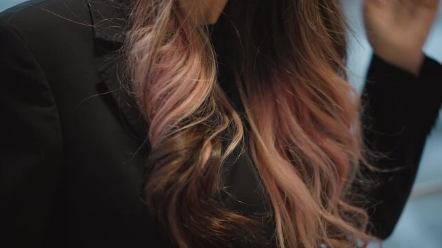 Waist-level close-up shows woman adjusting long wavy ombre hair with fingers, smoothing and twirling glossy chestnut to pink strands in soft window light for calm beauty and fashion moment