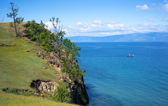 High angle view from a grassy cliff with trees overlooking Lake Baikal. Small boat sailing on deep blue water with mountains in the background. Summer nature landscape in Russia