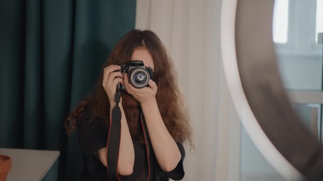 Curly-haired woman holds camera at eye level inside small studio, focusing on shot near window light and ring light, with hands on body and lens, face mostly hidden behind equipment