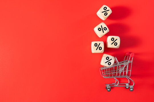 Miniature shopping cart surrounded by wooden percentage symbols on a vibrant red background, representing discounts, sales, savings, promotions, and retail shopping.