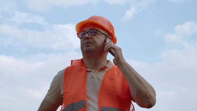 Construction worker in orange safety vest and hard hat standing outside talking on mobile phone. Worker receiving instructions from supervisor or foreman