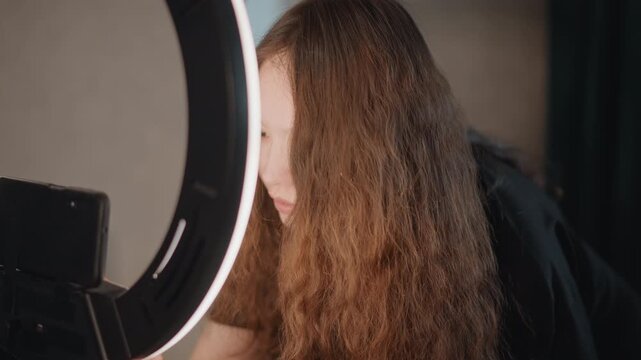 Young woman leans forward into illuminated LED ring light and adjusts smartphone mount with visible head tilt and hand movement, long wavy hair partially obscuring face