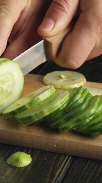 Close-up of hands slicing fresh cucumber into thin rounds on a wooden cutting board, showcasing the continuous action of cutting with a sharp knife in a well-lit kitchen setting
