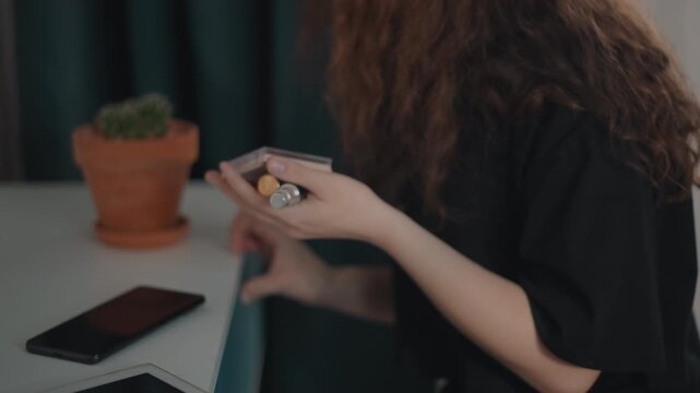 Close waist-level view of woman with long curly hair holding smartphone in right hand while left hand reaches toward terracotta potted succulent on table, capturing calm indoor lifestyle moment with