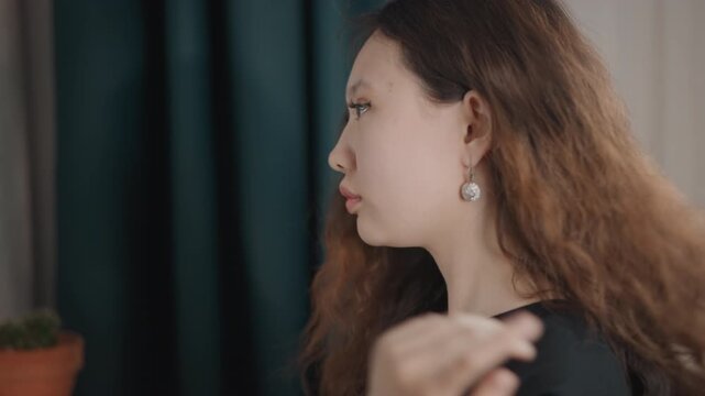 Profile closeup of young woman reaching up to adjust earring and tuck hair behind ear, with slight head turn, soft natural light, teal curtain backdrop, and calm grooming moment, with woman, profile