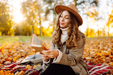 Fototapeta premium Woman in hat and coat sits on blanket in autumn park with book on sunny day. Beautiful woman reads book and relaxes surrounded by fallen leaves. Concept is autumn, relaxation, and enjoyment.