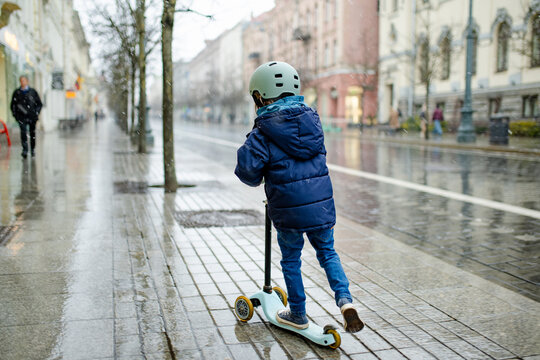Young child rides a kick scooter through the city during falling snow. Active outdoor childhood and urban transportation in cold winter weather.