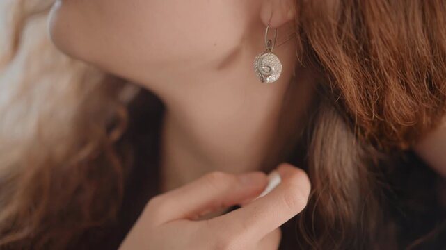 Profile view of young woman gently placing circular metallic earring into earlobe with thumb and forefinger, showing delicate hand movement, soft skin detail, warm light, and elegant beauty mood