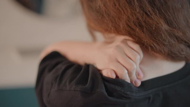Rear close-up of adult reaching over shoulder to press fingertips into upper back and neck base, with slight forward lean and hand contact suggesting self-massage, tension release, and quiet