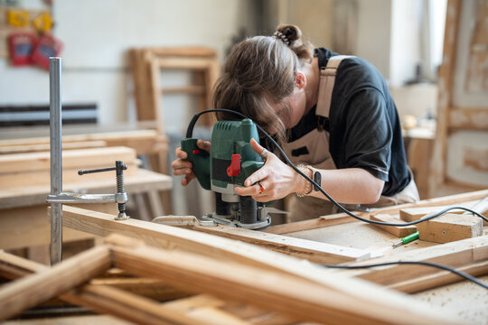 Woodworking restoration process in workshop, concentrated woman restorer using heavy wood router, female restorer cutting off wooden layers with electric plunge router, professional skills and craft.