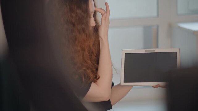 Seated person with long curly hair holds tablet toward camera and reaches to touch screen while turning head to side; face mostly hidden by hair, creating anonymity and reserved mood
