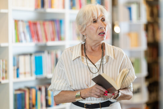 Focused mature lady browsing through literature volumes on shelves in cozy bookshop