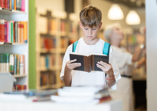 Interested teen male visitor holding a book near display stand in public library