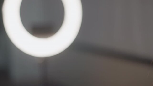 Bright circular ring light with phone mount on adjustable stand casting soft diffuse glow across nearby long dark brown hair at edge, in clean indoor beauty setting with gentle blur