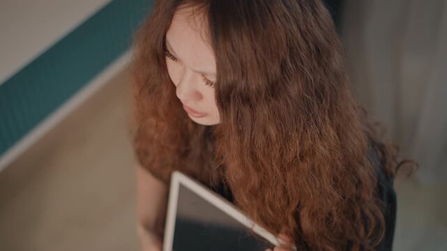 Top-down portrait of young woman with long curly hair lifting head slightly while holding notebook in soft indoor light, with subtle movement, reflective expression, calm focus and studious mood