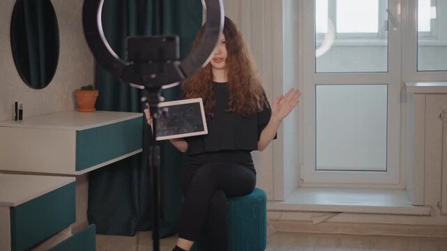 Young woman sits on upholstered stool holding tablet while speaking and gesturing toward camera beside glowing ring light in bright modern interior with soft daylight and clean home studio styling