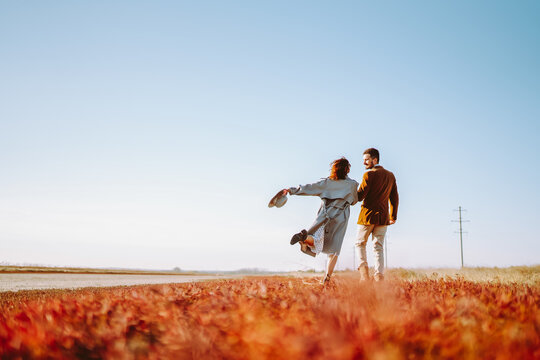 Young man and woman walk together across bright grassy field at sunset. A loving couple in stylish clothing enjoys nature and time together outdoors. Concept of love and relaxation.