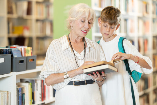 Happy senior woman and boy choosing books near high wooden shelves in bookstore aisle