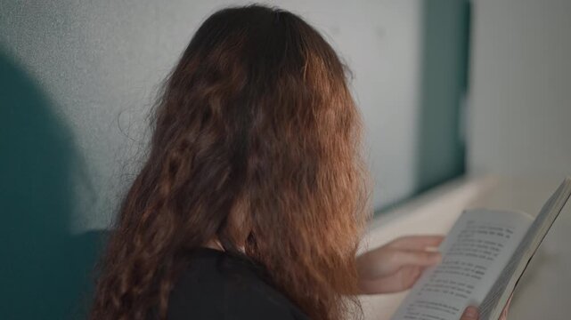 Close over shoulder view of young woman with long curly hair turning pages of booklet, hands on paper while reading with focused expression, teal wall and soft side light creating calm indoor mood