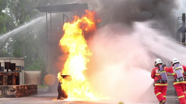 Firefighter fire hose water foam at industrial plant, emergency rescue training drill, protective crew spraying powerful stream on fuel tank blaze, thick smoke, safety practice