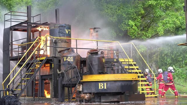 Firefighter fire training at industrial facility, water hose stream sprays burning tank platform, refinery drill with smoke and flame, emergency response crews control hazard outdoors
