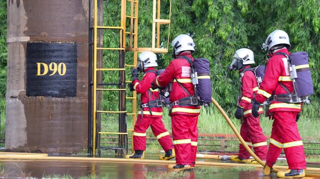 Firefighter fire hose water spray emergency response safety training drill at industrial tank site, team in protective gear with breathing apparatus, helmet and ladder, controlling jet near forest