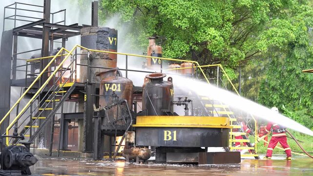 Firefighter hose water spray industrial plant tank pipeline safety training emergency response outdoor firefighting drill, crew in protective gear controlling smoke, high pressure jet cooling