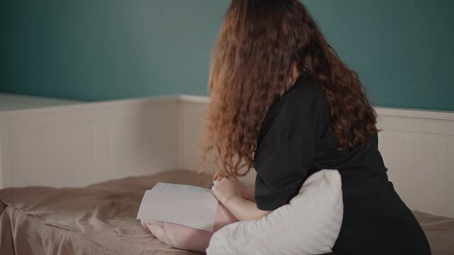 Young woman sits on bed leaning forward while hugging white textured pillow, hair covering face and body curled inward in quiet bedroom setting with soft natural light and reflective mood