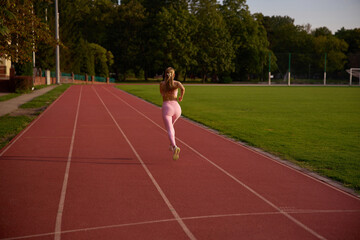 Obraz premium Female runner in motion during beautiful golden hour sunlight. Active outdoor fitness, physical exercise, and athletic wellness concept. Young woman sprinting on a sports track