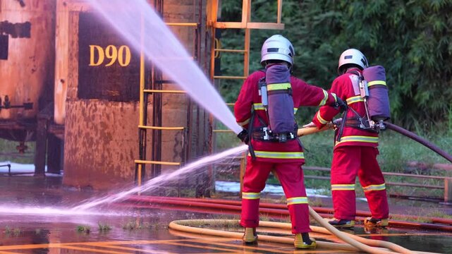 Firefighter with fire hose and water spray industrial emergency safety training drill, protective gear teamwork directs high pressure stream toward factory storage tank, crew extinguish blaze wet
