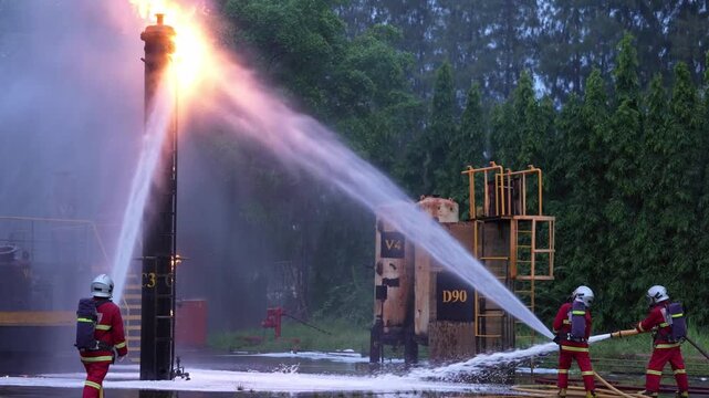 Firefighter fire hose water stream battling industrial flame during emergency training safety drill, protective gear crew spraying pressure jet to control refinery fuel pipe blaze outdoors dusk