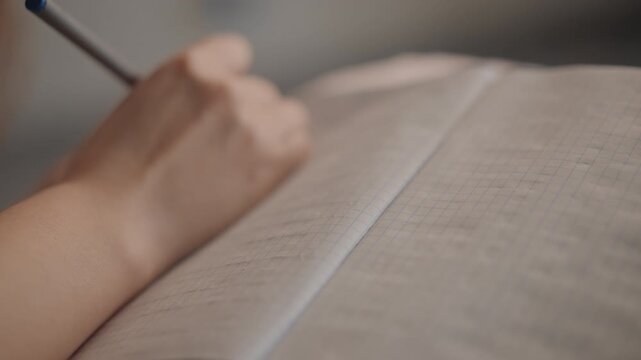 Over shoulder close view of hand holding pencil above open book, fingers relaxed in brief pause before writing, with warm light, shallow focus, and calm indoor workspace atmosphere