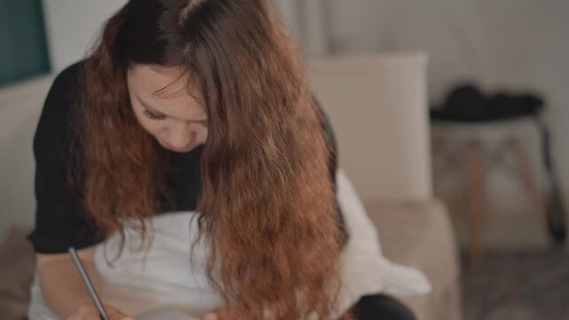 Young woman seated on bed leans forward while writing in notebook, turning page with one hand and holding pen ready to continue, hair falling across face under warm soft indoor light in calm bedroom