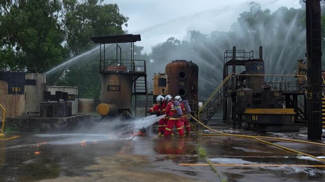 Firefighter fire drill at industrial plant refinery yard, emergency response team using hose streams, water spray and foam to suppress burning fuel spill, heavy smoke mist, safety training exercise