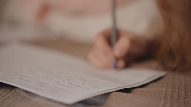Over shoulder close-up of hand holding pen and writing on paper in warm indoor light, visible pen tip movement, soft blur, focused study mood, desk surface, casual sleeve, and intimate indoor setting