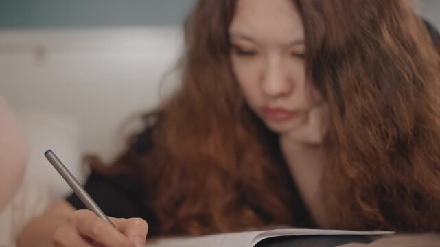Close eye-level view shows young person writing in lined notebook with pen, hand moving across paper during active note taking beside desk items under warm soft indoor light, with partial face and