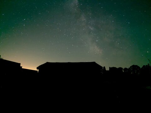 Voie lact&eacute;e visible dans un ciel &eacute;toil&eacute; d'&eacute;t&eacute; au-dessus de silhouettes de b&acirc;timents et d'arbres &agrave; la campagne, photographi&eacute;e de nuit en Bretagne pendant la p&eacute;riode des Pers&eacute;ides au mo