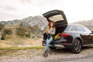 Fototapeta premium Traveler wearing hat and holding phone sits in trunk of car overlooking mountains. Woman on road trip enjoys mountain scenery with phone. Concepts of adventure, transportation, and freedom.