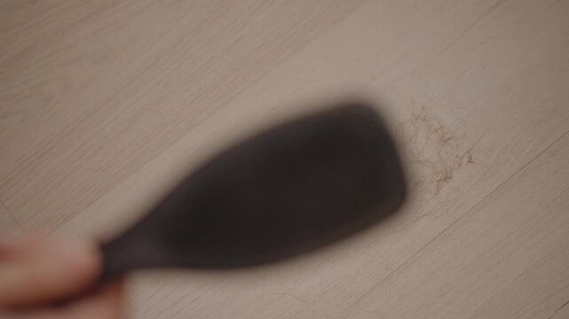 Medium-close low angle view shows woman holding dark hairbrush above pale wooden or laminate surface, with gentle hand movement, visible brush teeth, soft blur, and subtle residue in clean studio