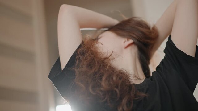 Close portrait of young woman styling long curly hair while raising arms, tilting head, and leaning forward in soft indoor light, with flowing strands, calm expression, and graceful personal grooming